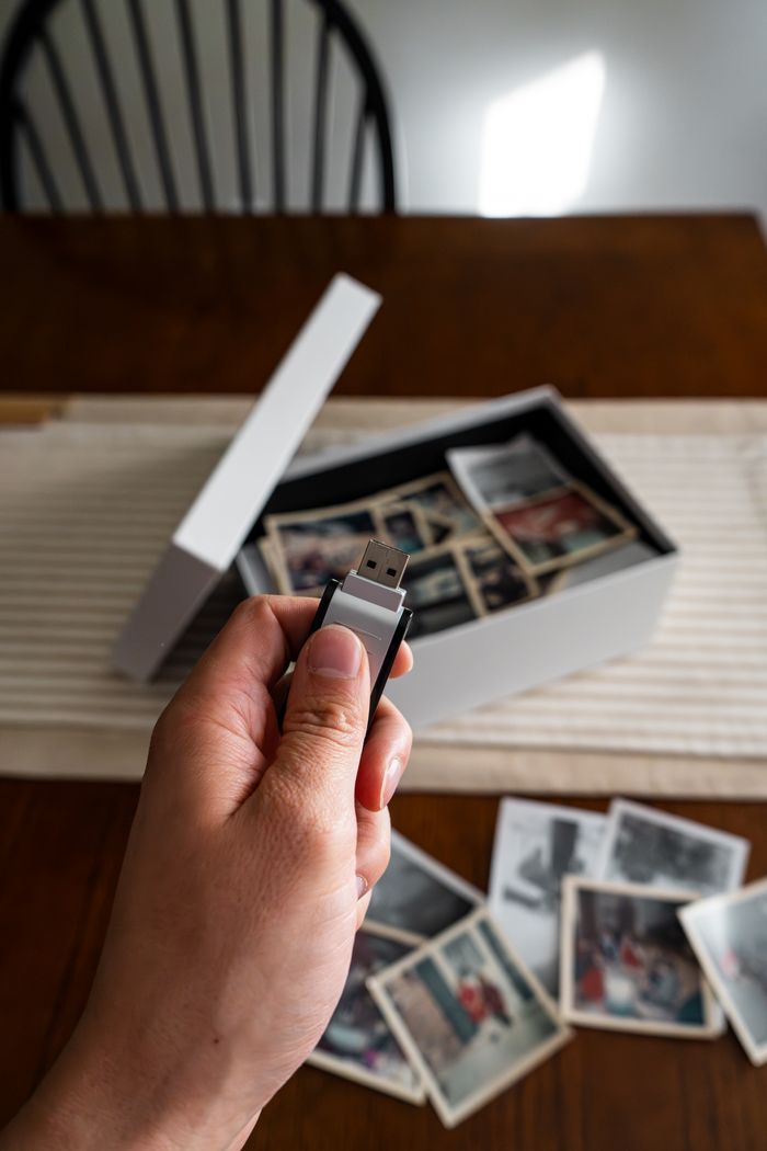 Hand holding USB drive above box of old family photos on a table.